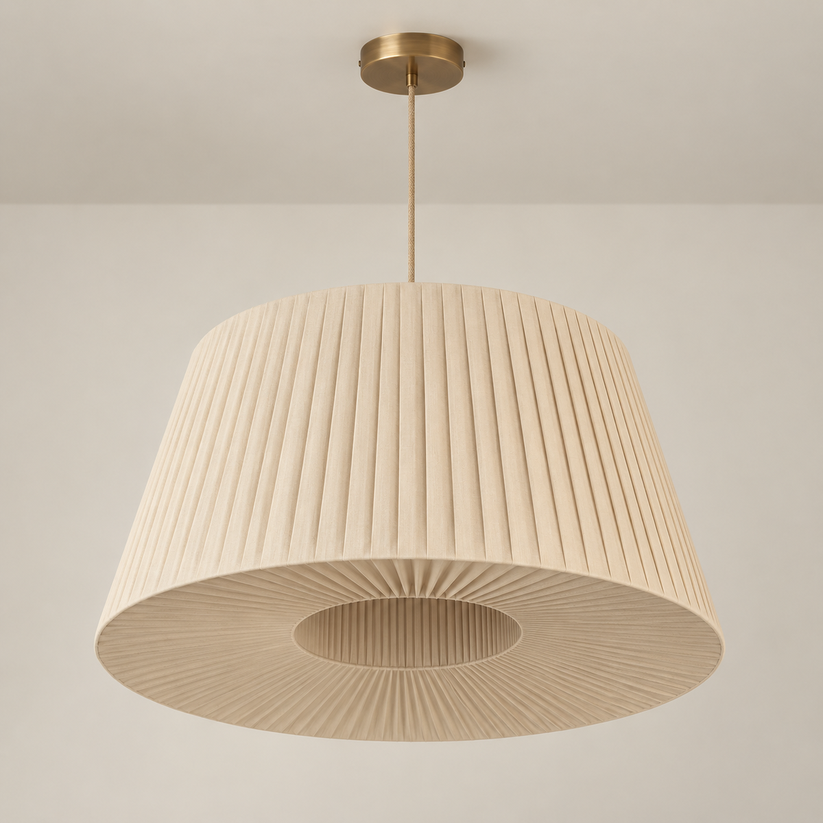 A modern pleated drum pendant light in a warm beige fabric shade with a brushed brass ceiling canopy and rod, shown hanging against a neutral off-white wall.