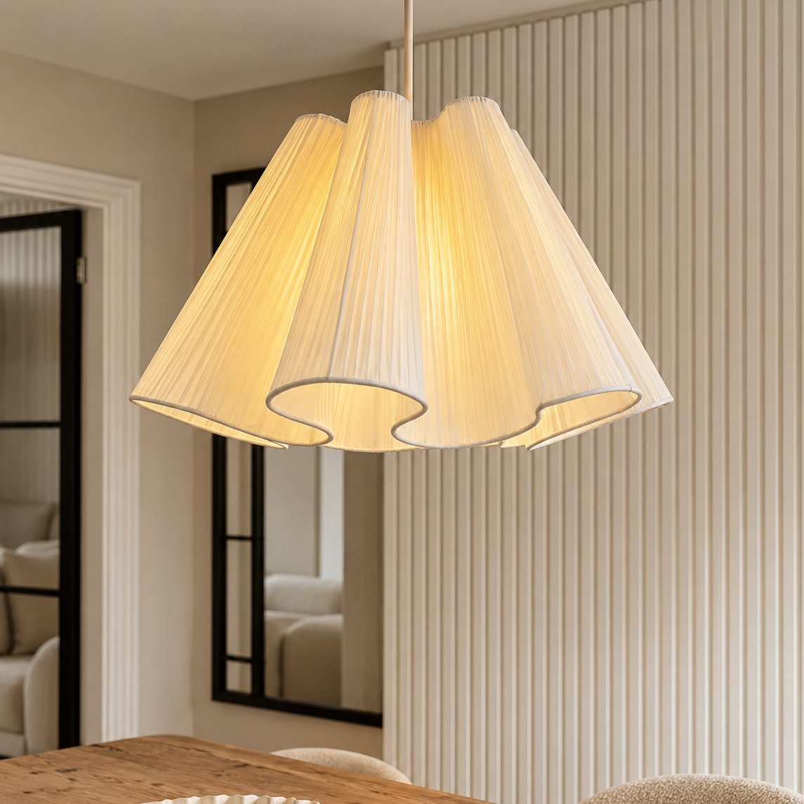 A modern pleated fabric pendant light in a warm cream color, hanging over a wooden dining table in a stylishly decorated room with beige walls and a slatted accent wall.