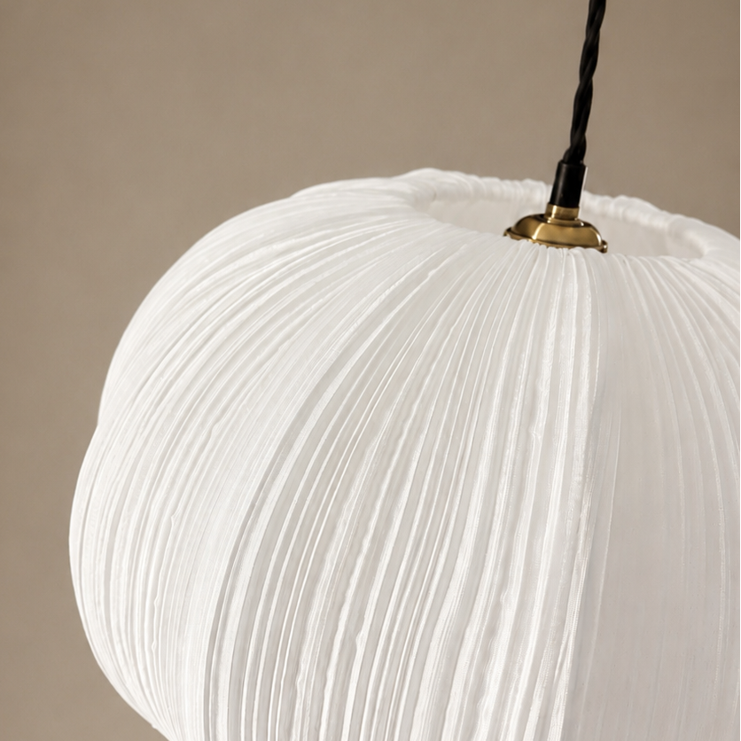 A close-up of a white pleated fabric pendant light shade hanging from a black cord with a small gold metal cap, set against a solid warm beige background.