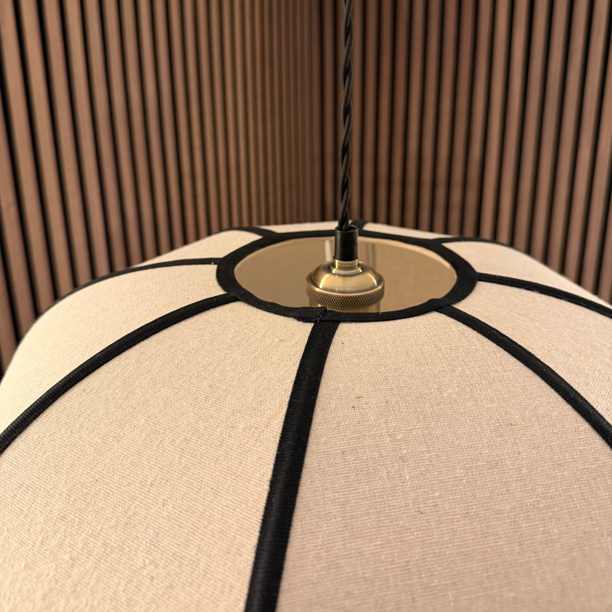Close-up of a hanging pendant lamp with a cream fabric shade, black cord trim, and a brass metal fixture, suspended in front of brown wood slat walls.