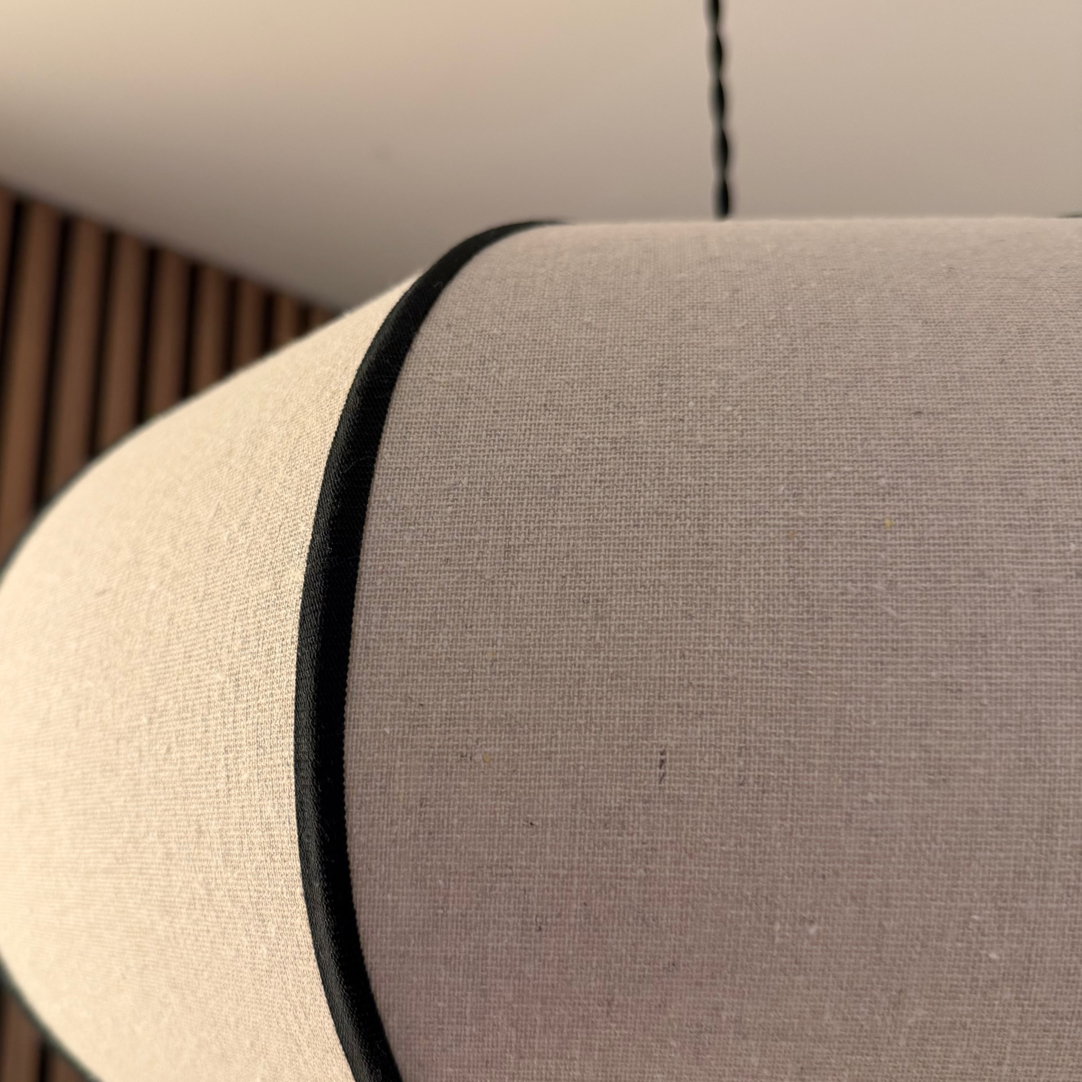 A close-up of a cylindrical pendant light shade made of light beige textured fabric, featuring a thin black trim band and a black hanging cord against a modern slatted wood wall background.