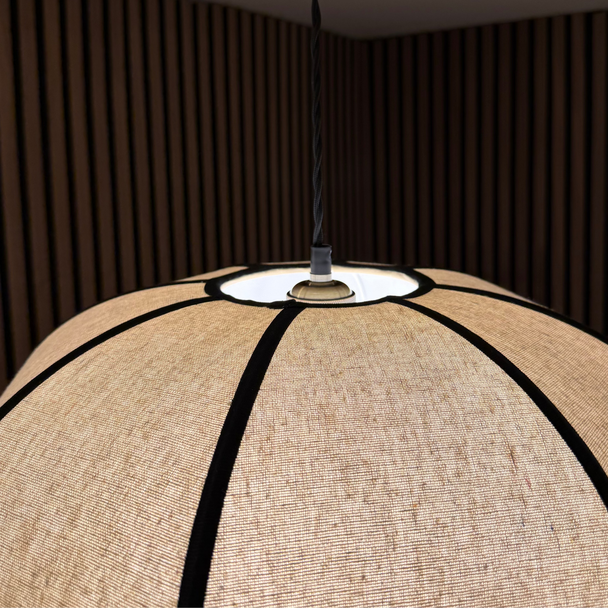 A close-up of a large spherical pendant lampshade made of beige woven fabric with black curved trim, suspended by a black cord in a room with dark wood slat walls.