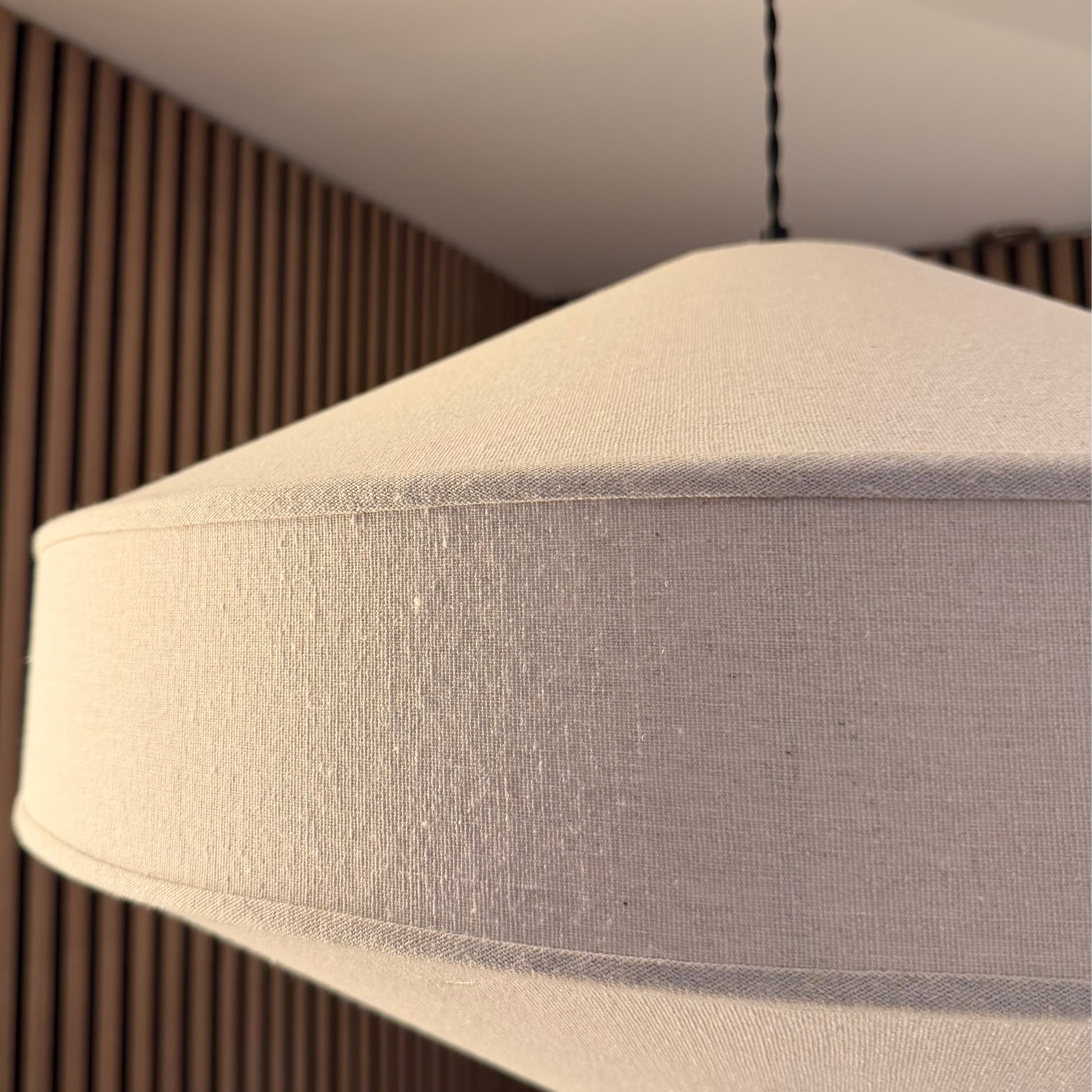 A close-up of a modern pendant light with a textured off-white fabric shade, hanging in a room with a slatted wood wall background.