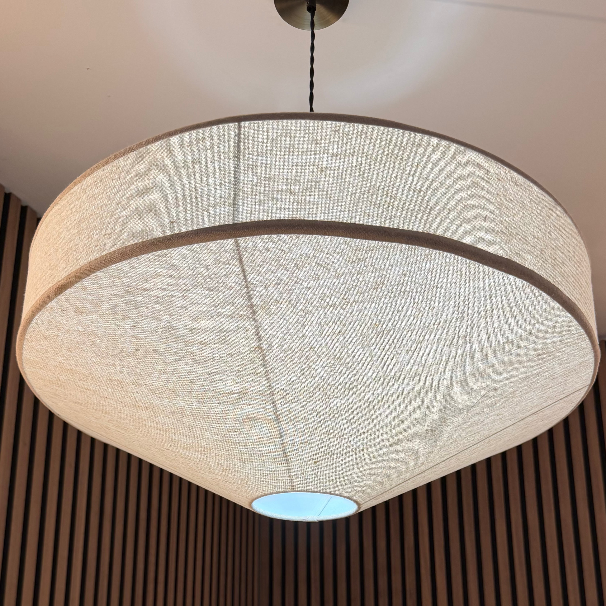A large modern drum pendant light with a textured beige linen shade hanging in a room with dark brown vertical wood slats and a green plant below.