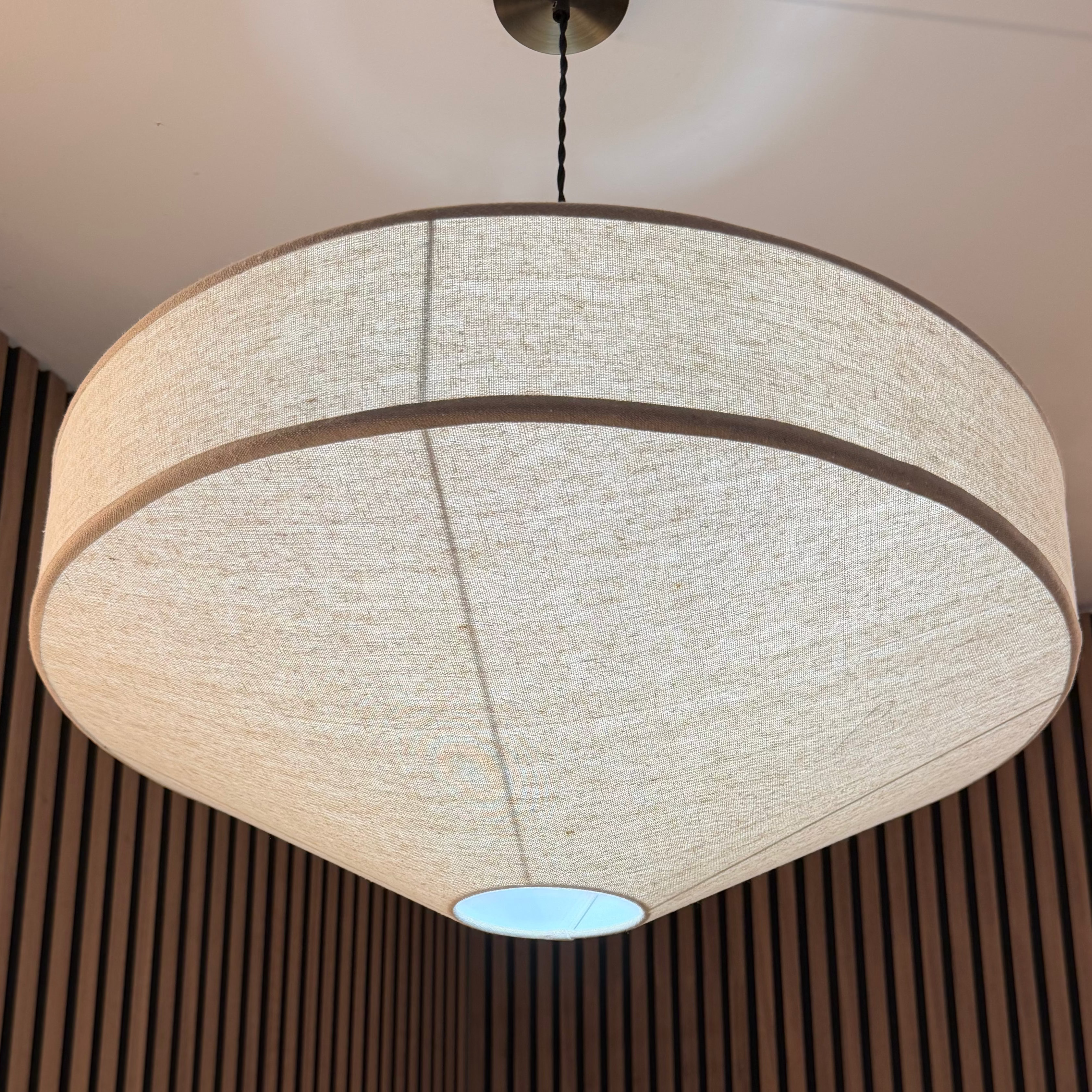 A large modern drum pendant light with a textured beige linen shade hanging in a room with dark brown vertical wood slats and a green plant below.