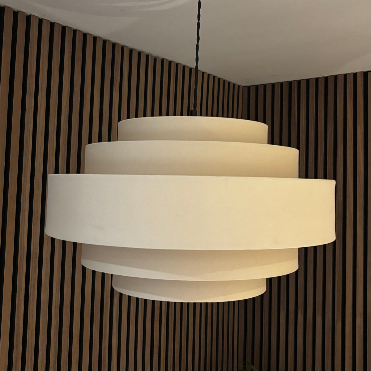 A modern three-tier tiered pendant chandelier with a warm off-white shade, suspended from a brass ceiling canopy against a brown wood slat wall.