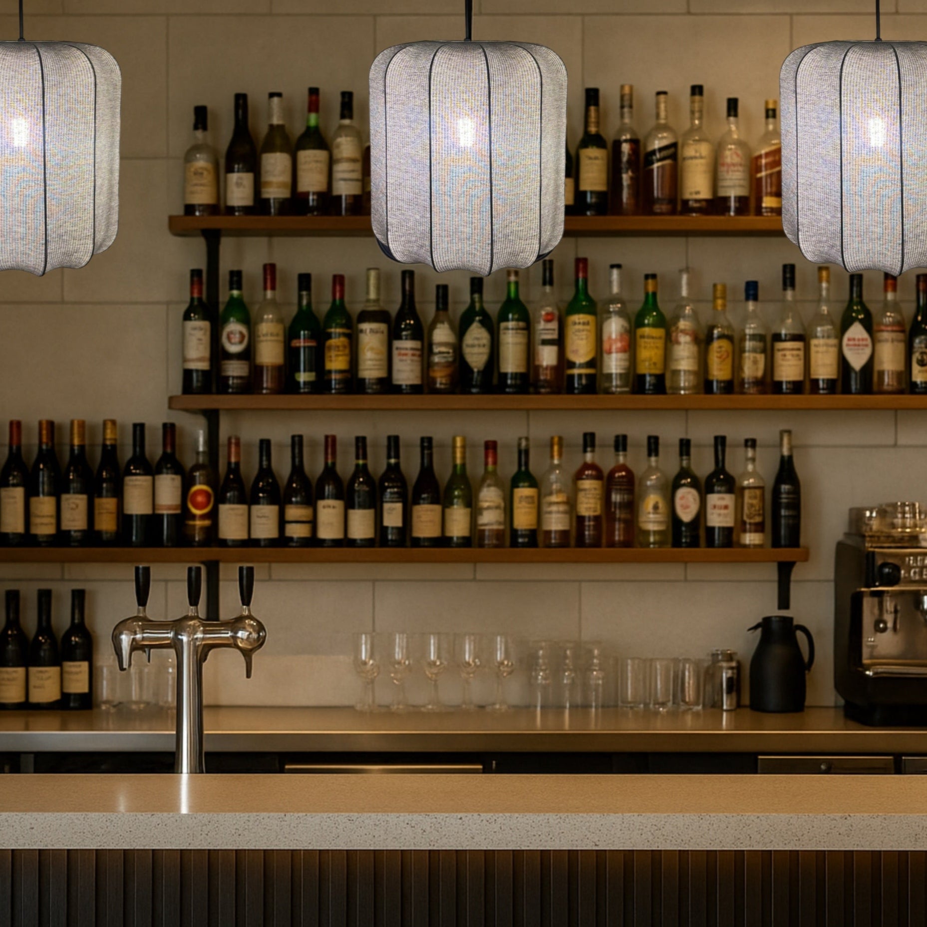 Bar setup with shelves of alcohol bottles, glasses, and a coffee machine under pendant lights.