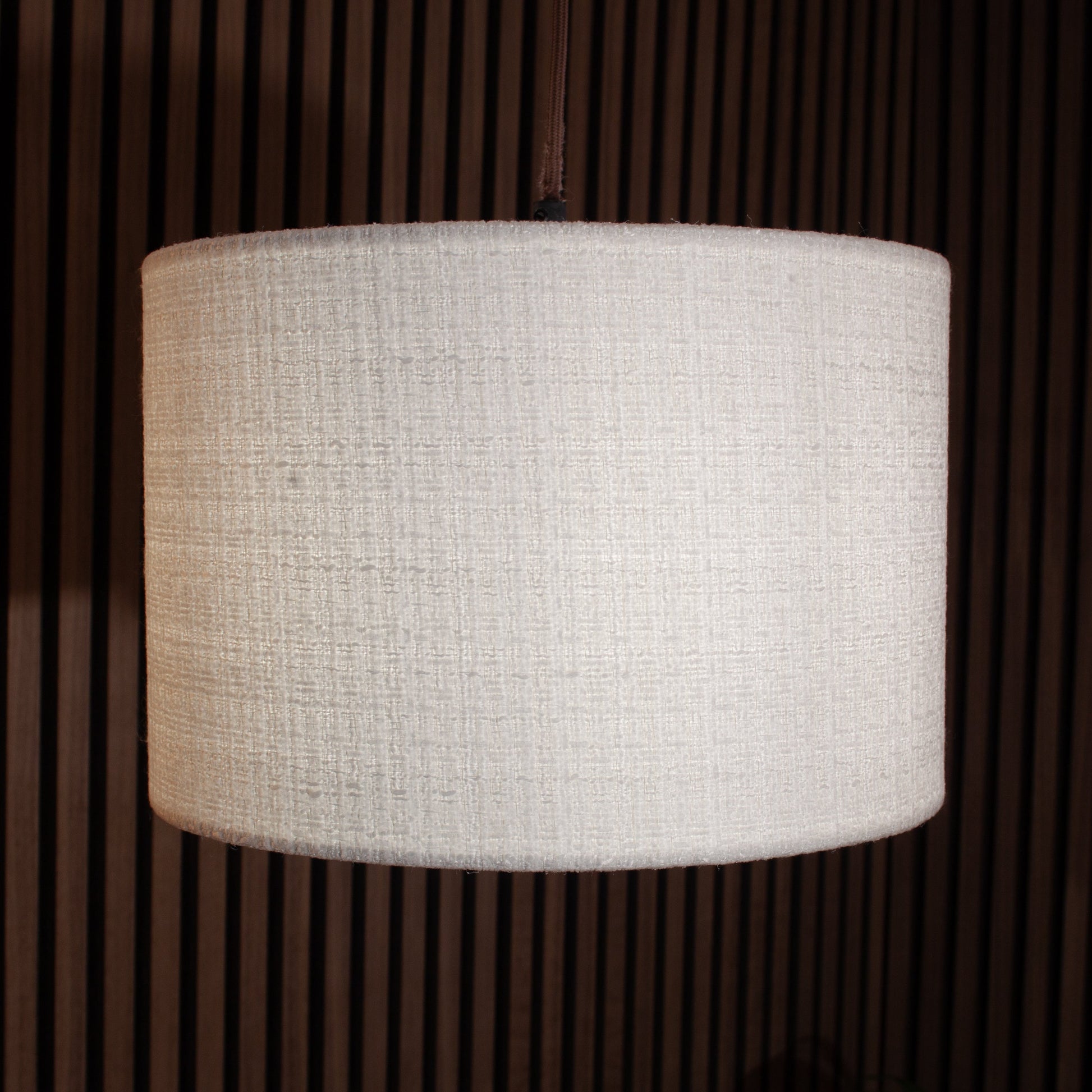 A cylindrical drum lampshade made of textured off-white fabric, shown hanging against a dark brown slatted wooden background.