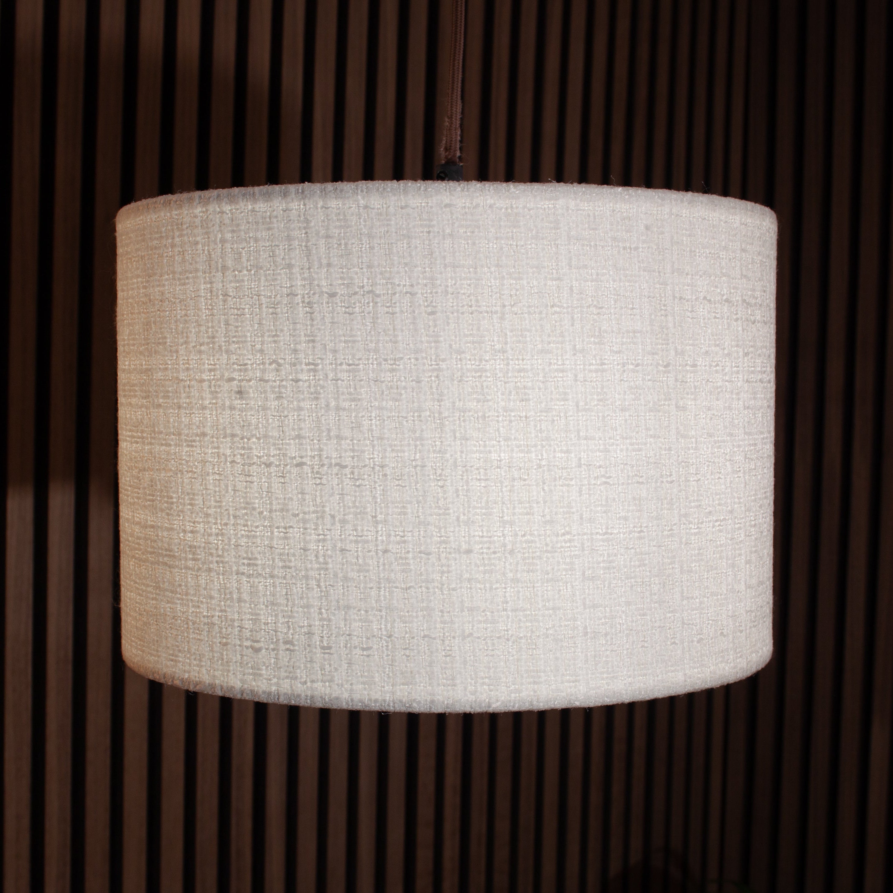 A cylindrical drum lampshade made of textured off-white fabric, shown hanging against a dark brown slatted wooden background.