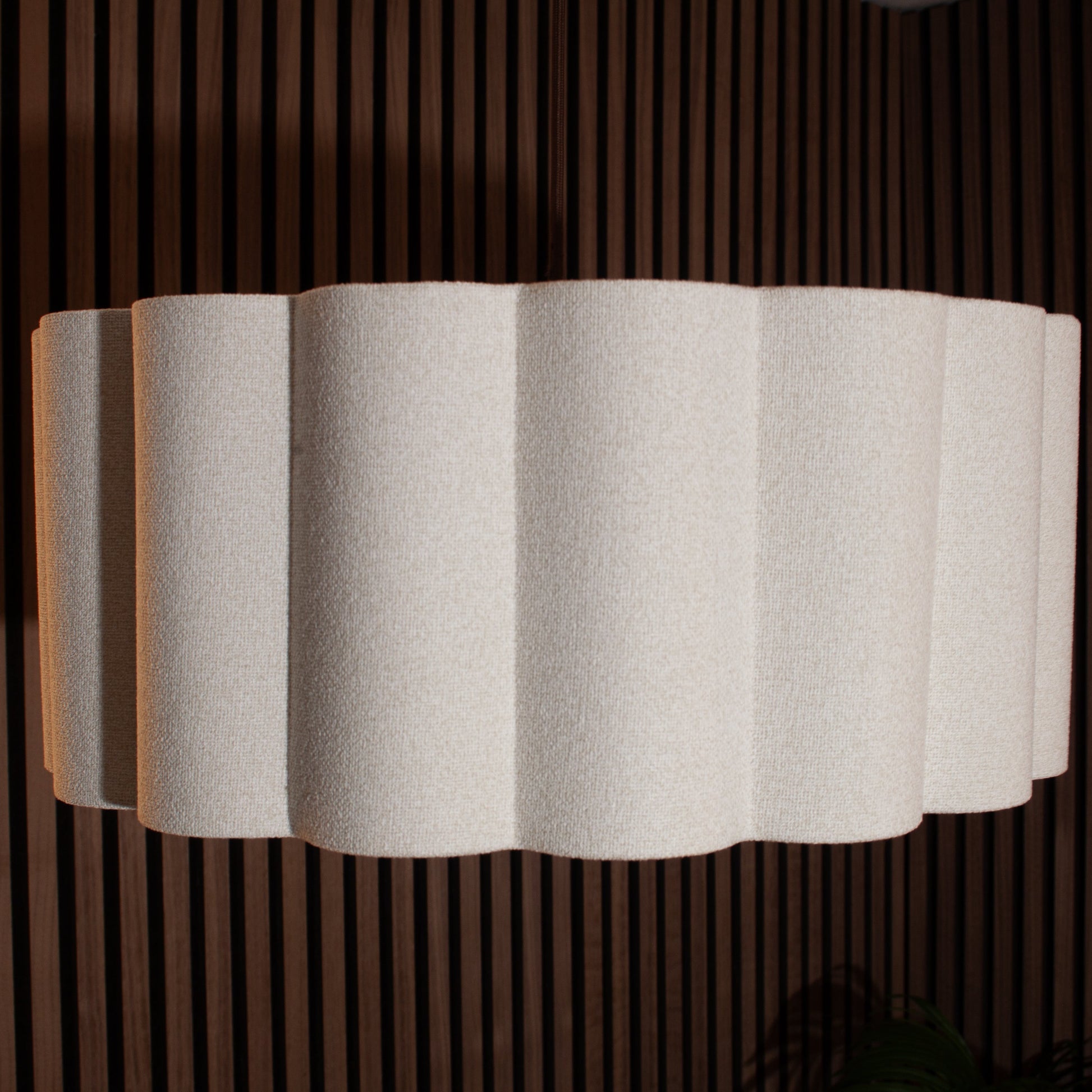A modern wall sconce lamp featuring a pleated, scalloped white fabric shade, mounted against a dark brown vertical slat wood screen.