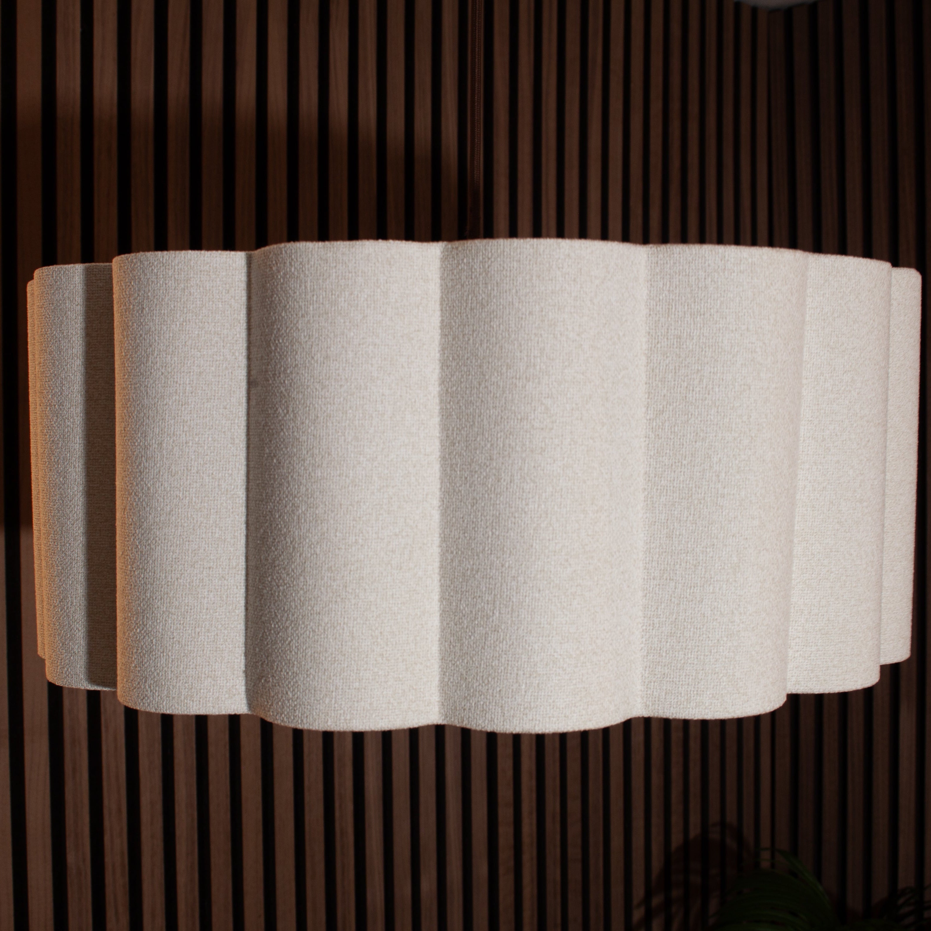 A modern wall sconce lamp featuring a pleated, scalloped white fabric shade, mounted against a dark brown vertical slat wood screen.