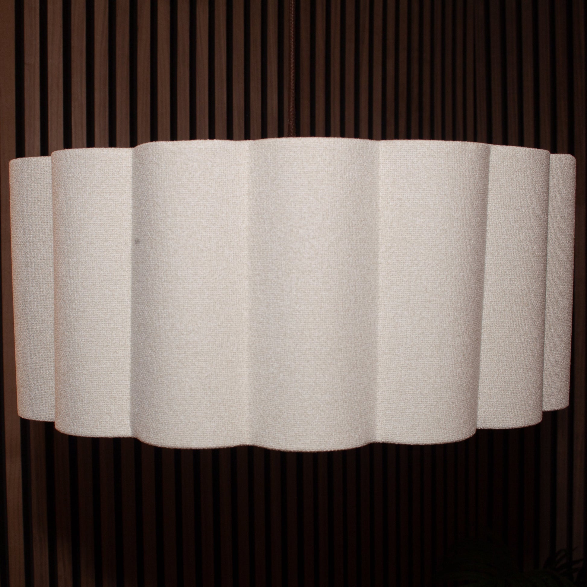 A modern wall sconce featuring a wavy, pleated white fabric shade mounted against a dark brown vertically slatted wooden wall.
