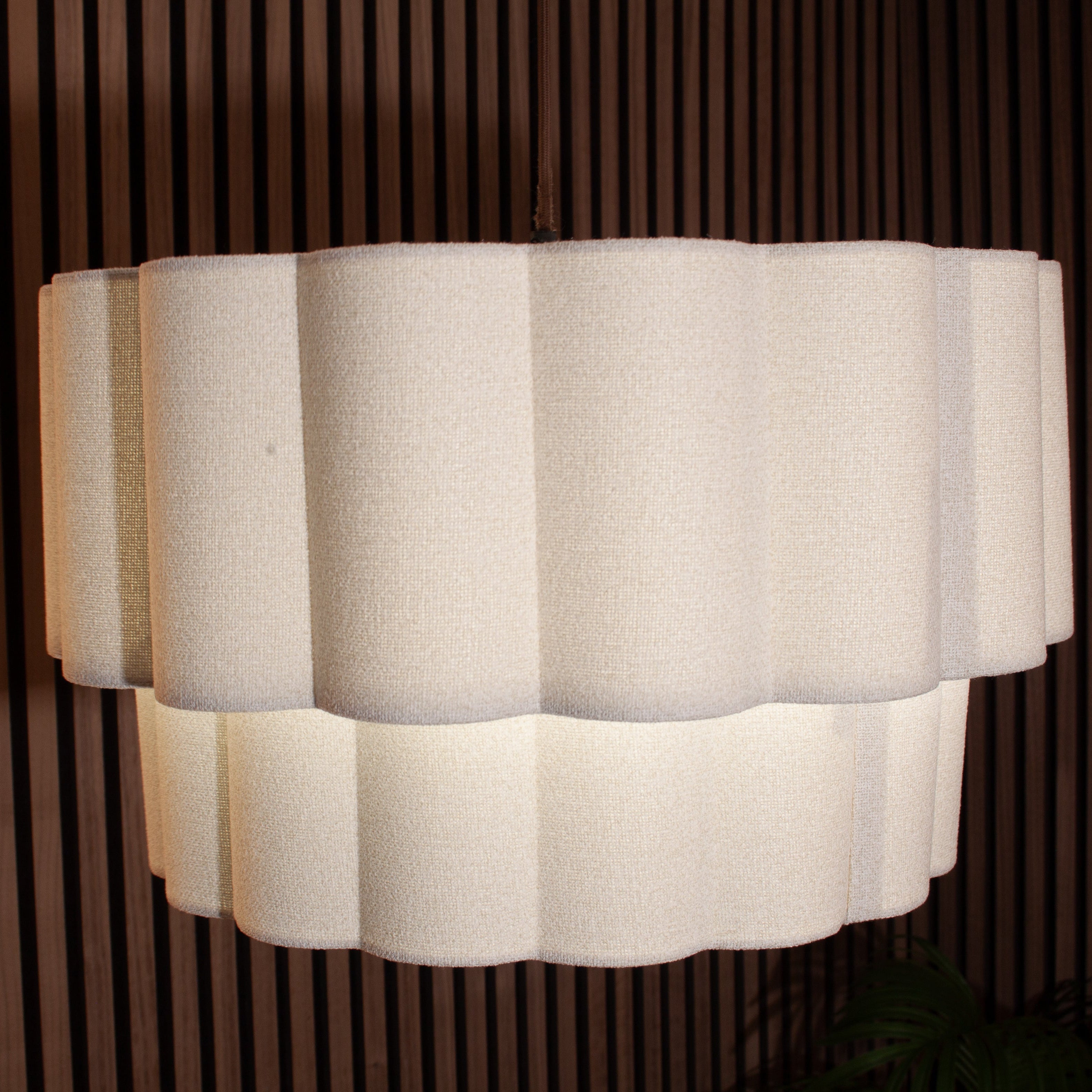 A modern tiered drum pendant light with a textured off-white fabric shade, shown illuminated against a dark brown vertical slat wall.