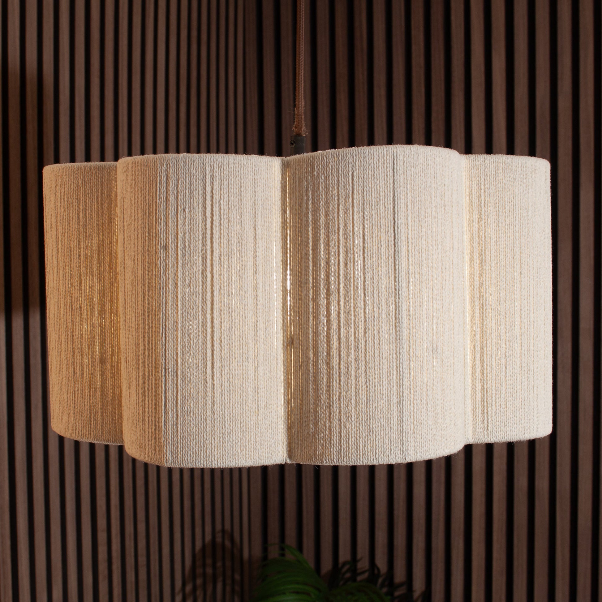 A modern multi-tiered pendant light with a textured, pleated beige fabric shade, suspended against a dark brown slatted wood wall.