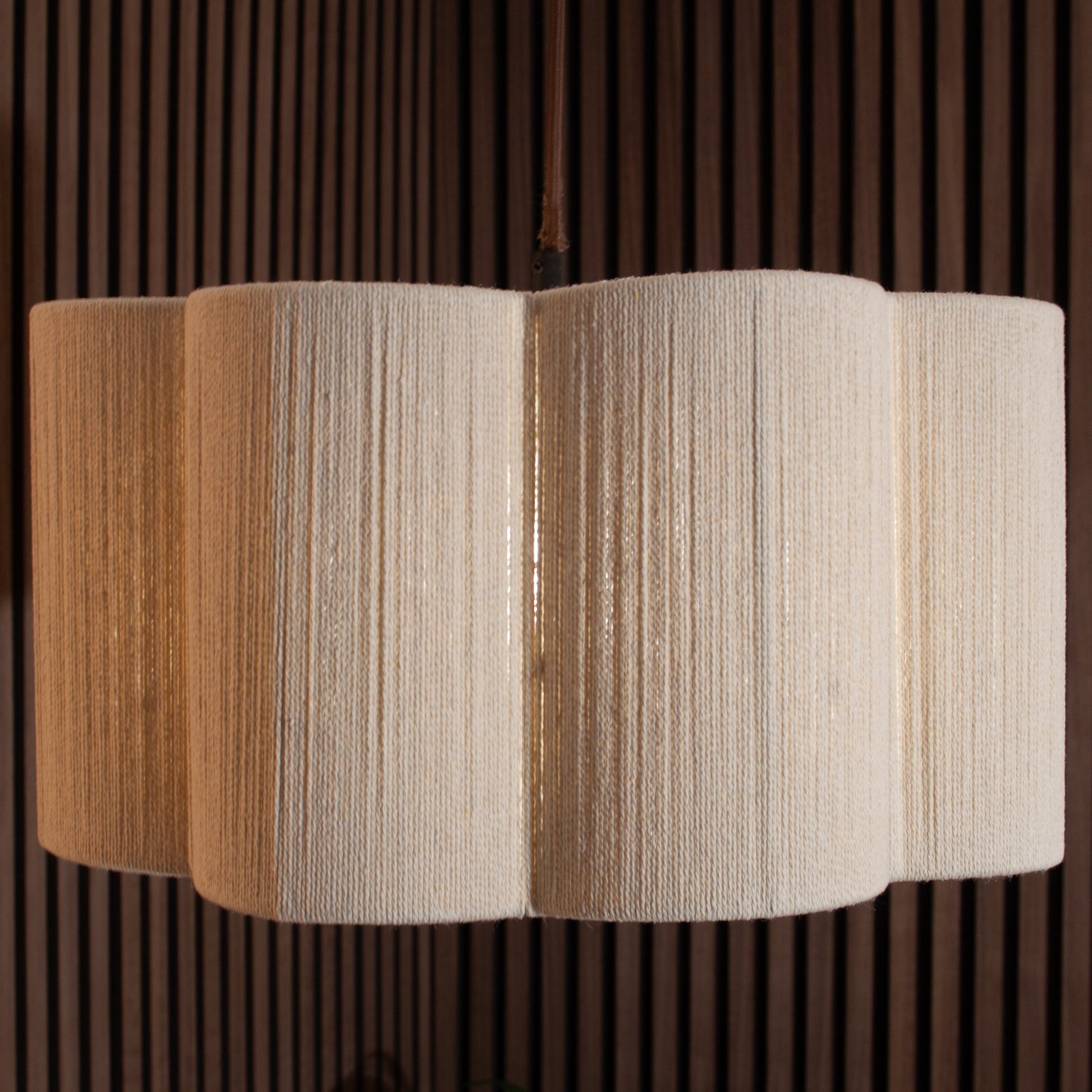 A modern multi-tiered pendant light with textured cream fabric shades, suspended against a dark brown slatted wood wall.