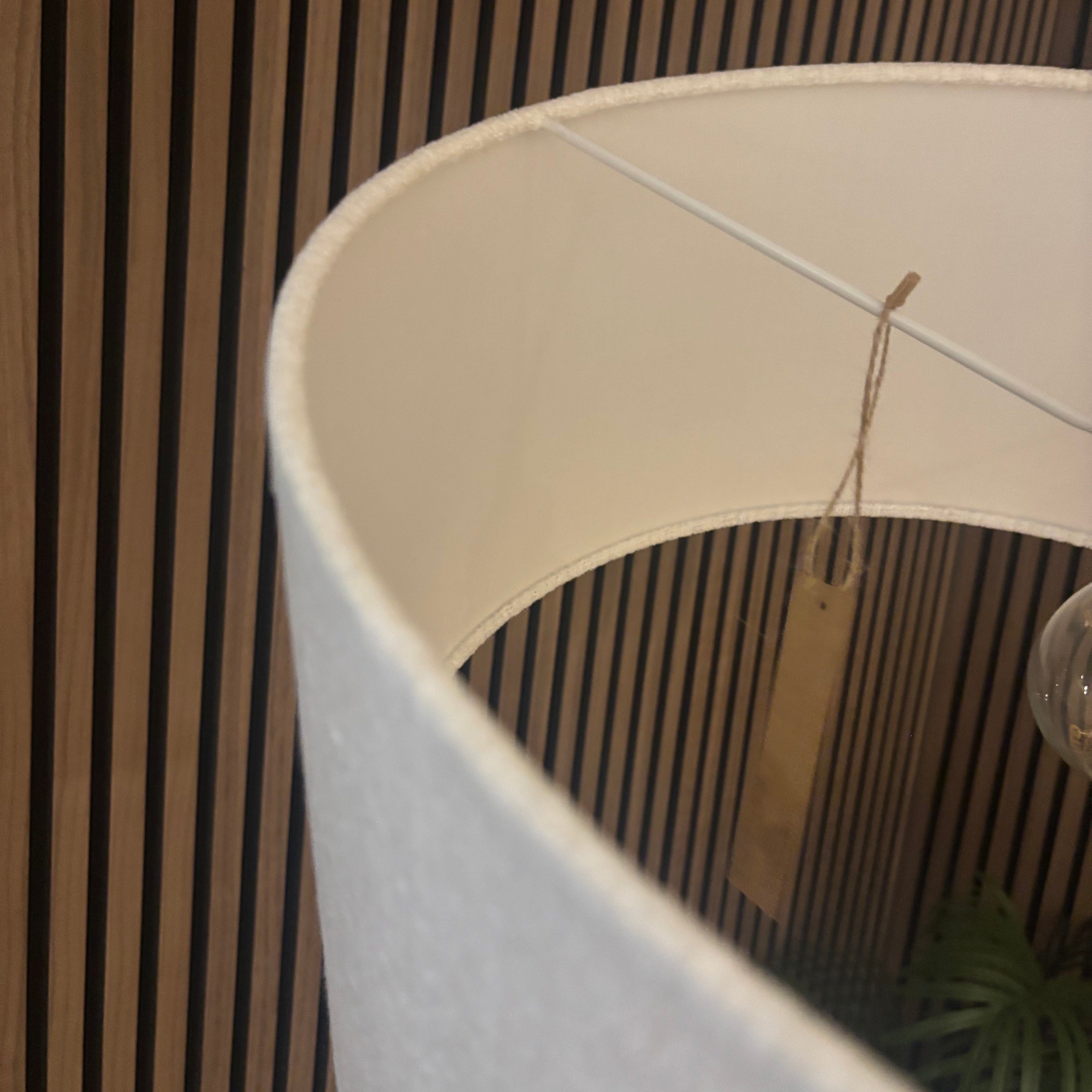 Close-up of a large white fabric lampshade with a hanging kraft paper tag, set against a dark wood slat wall.
