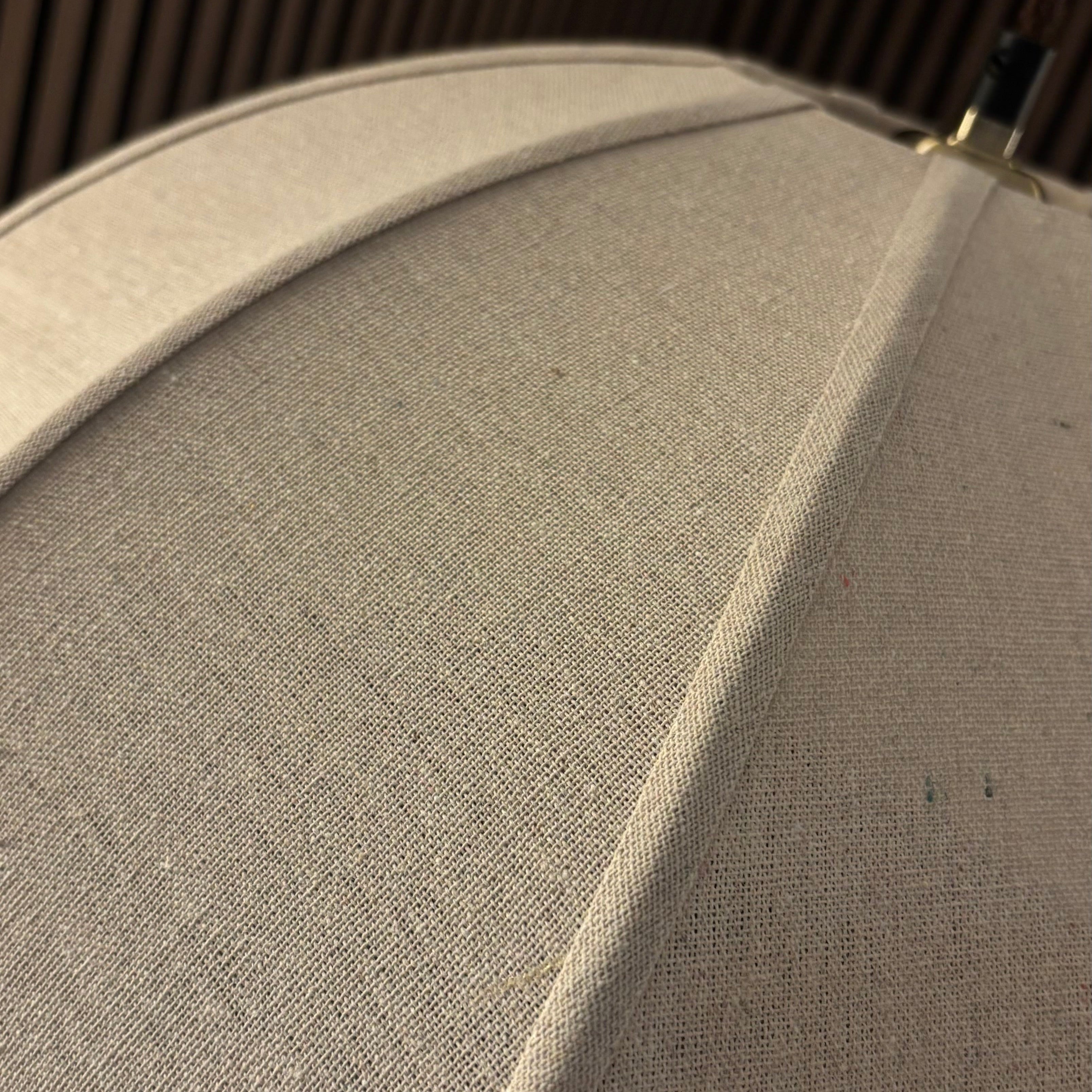 Close-up of a light beige fabric lampshade with visible woven texture and a dark metal finial at the top, set against a dark slatted background.