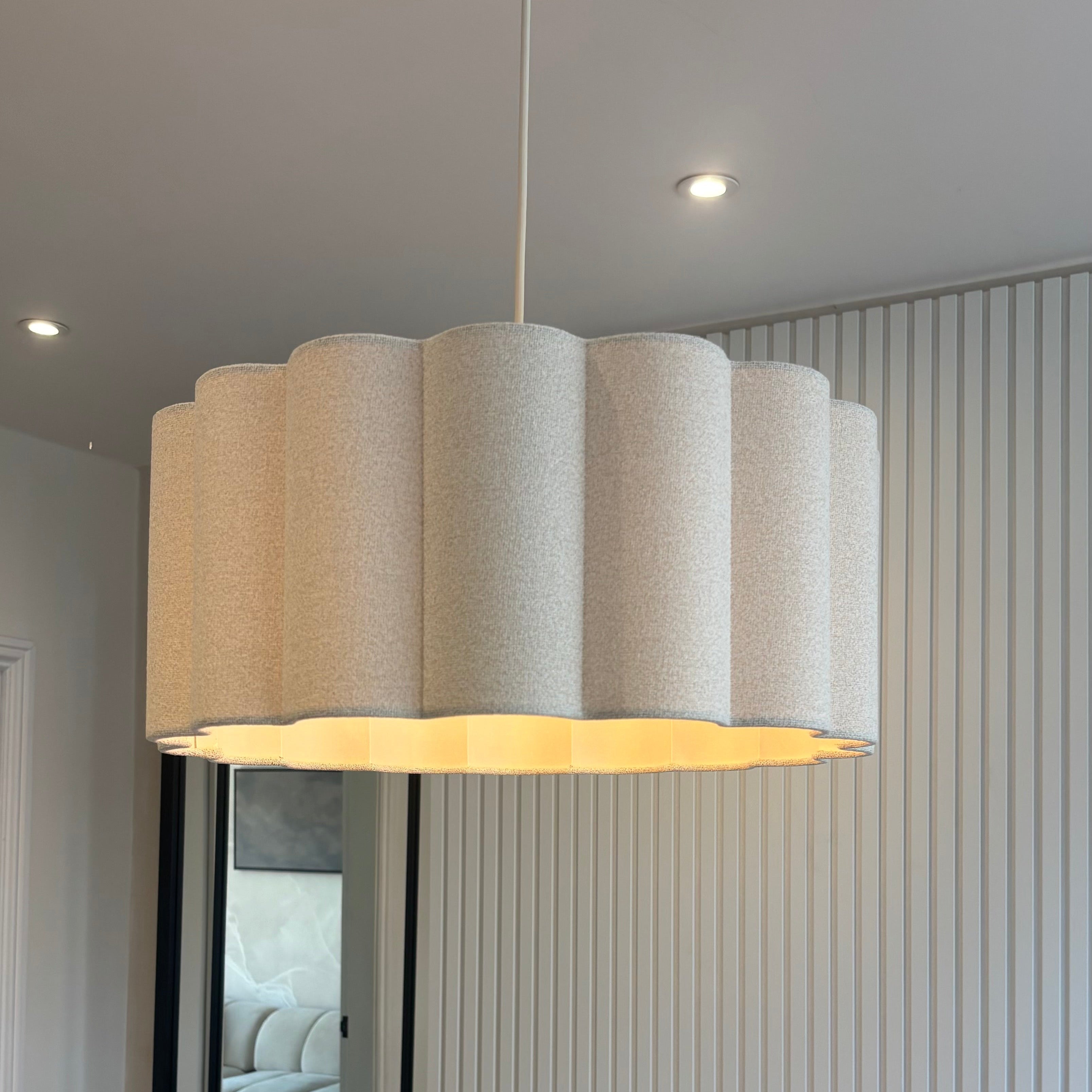 A modern pendant light with a scalloped drum shade made of textured cream fabric, hanging in a contemporary room with a ribbed white wall and a wooden table in the foreground.