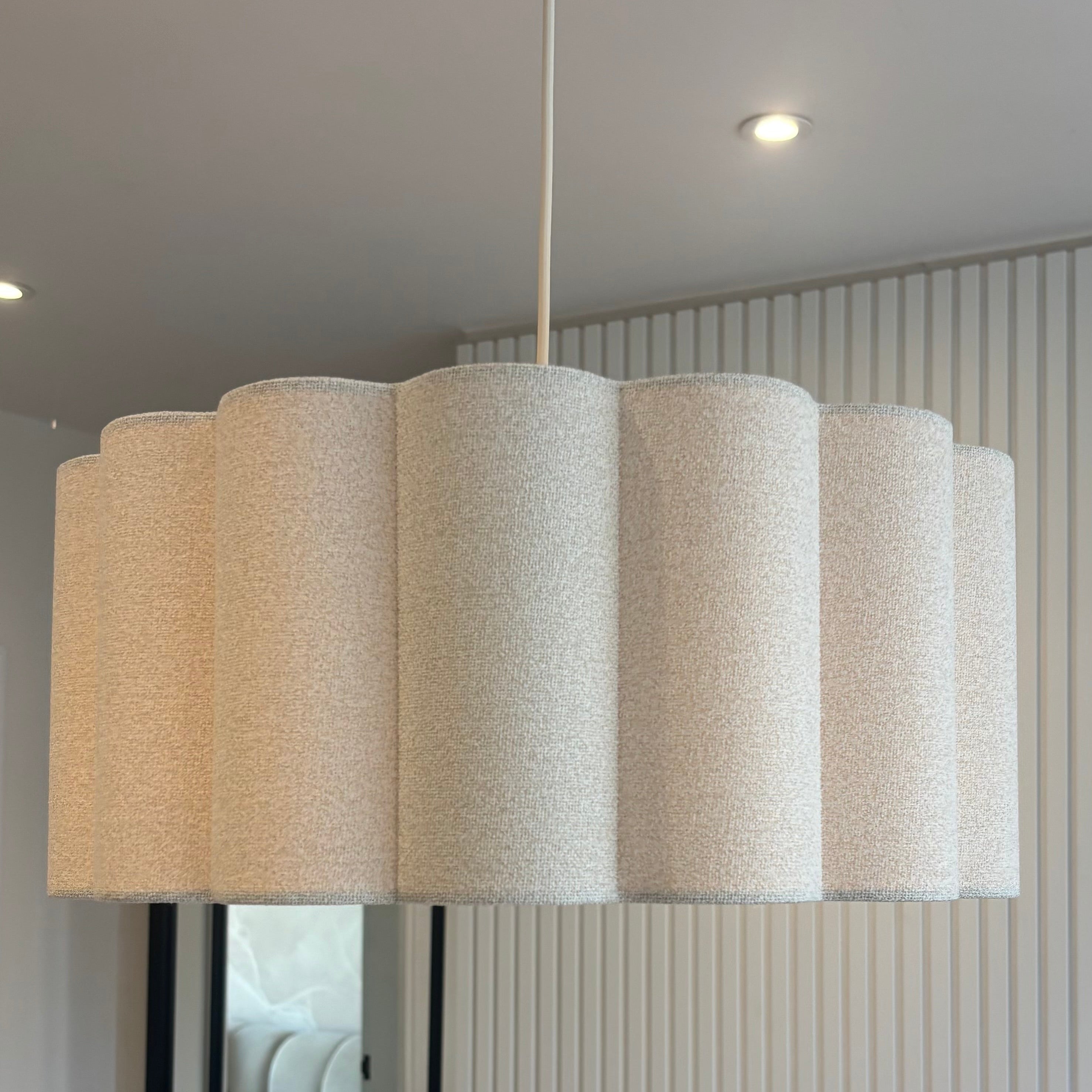A modern pendant light with a series of fluted cylindrical shades in a textured cream fabric, hanging in a contemporary room with a ribbed white wall and a mirror.