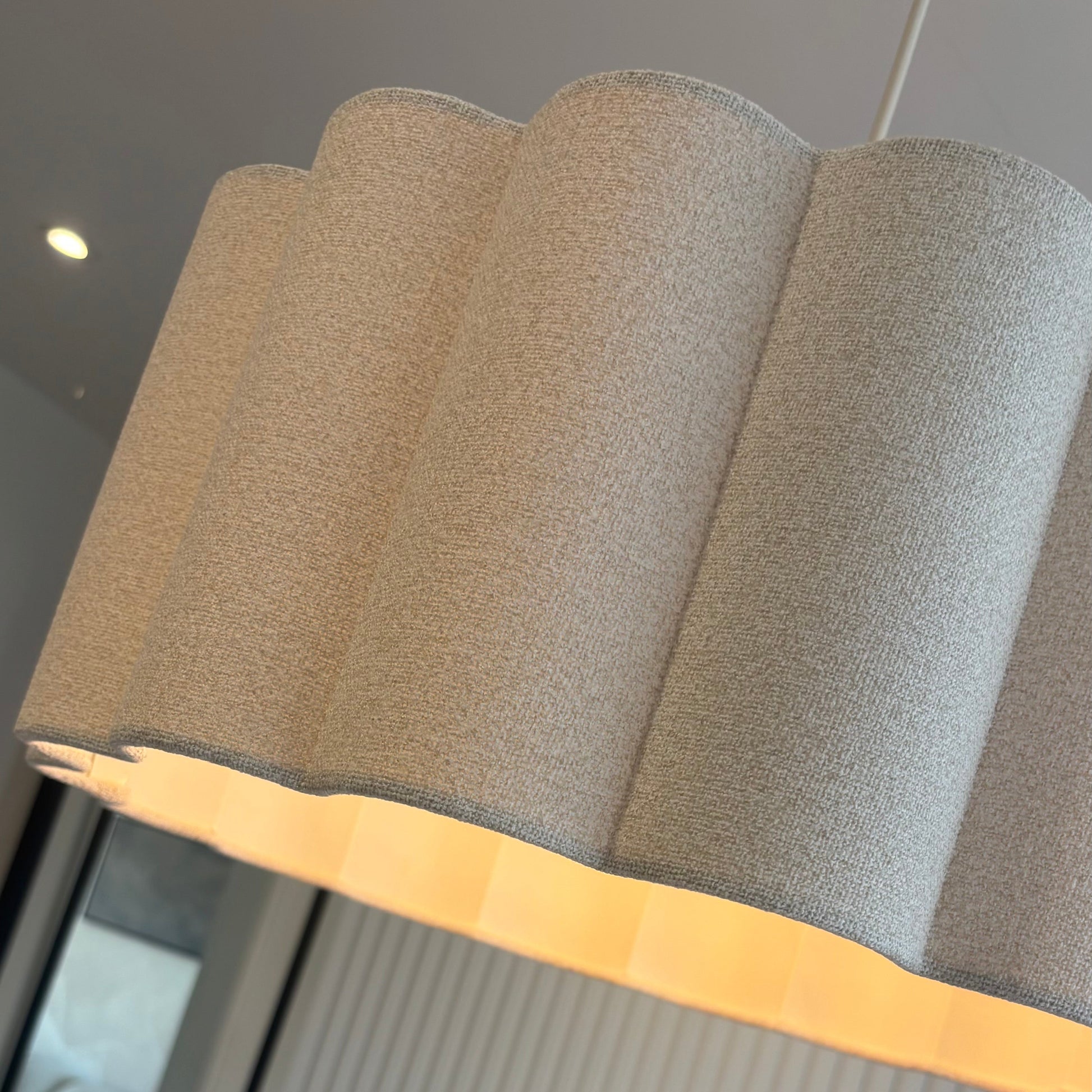 A close-up of a modern pendant light with a textured beige fabric shade featuring a scalloped or wavy fluted edge and a warm yellow-gold inner trim, shown hanging in an interior setting.