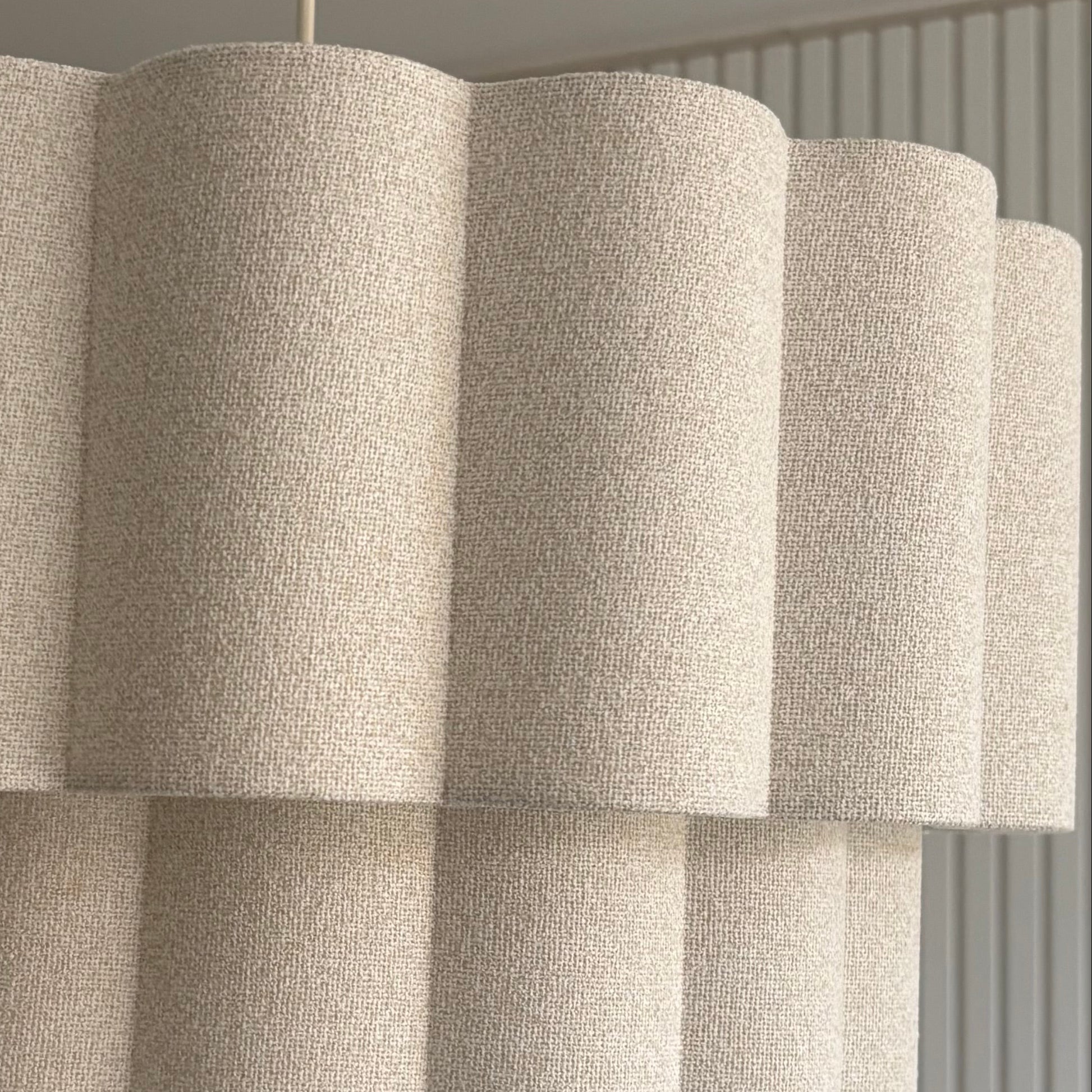 A modern multi-tiered chandelier pendant light with textured beige fabric shades, hanging in a contemporary room with a ribbed white wall and a mirror.