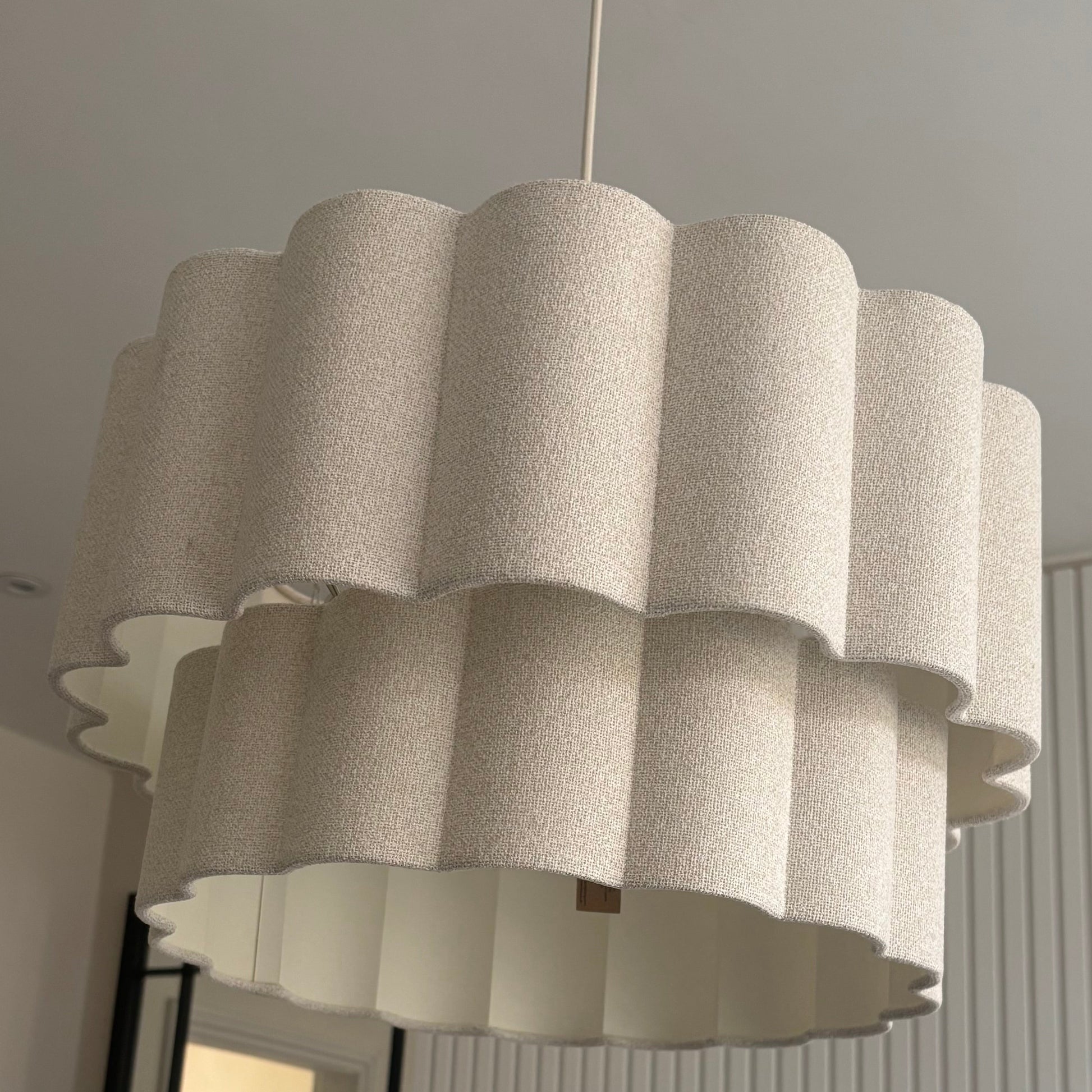 A modern tiered chandelier pendant light with a textured cream fabric shade, hanging in a contemporary room with a slatted white wall and a mirror.