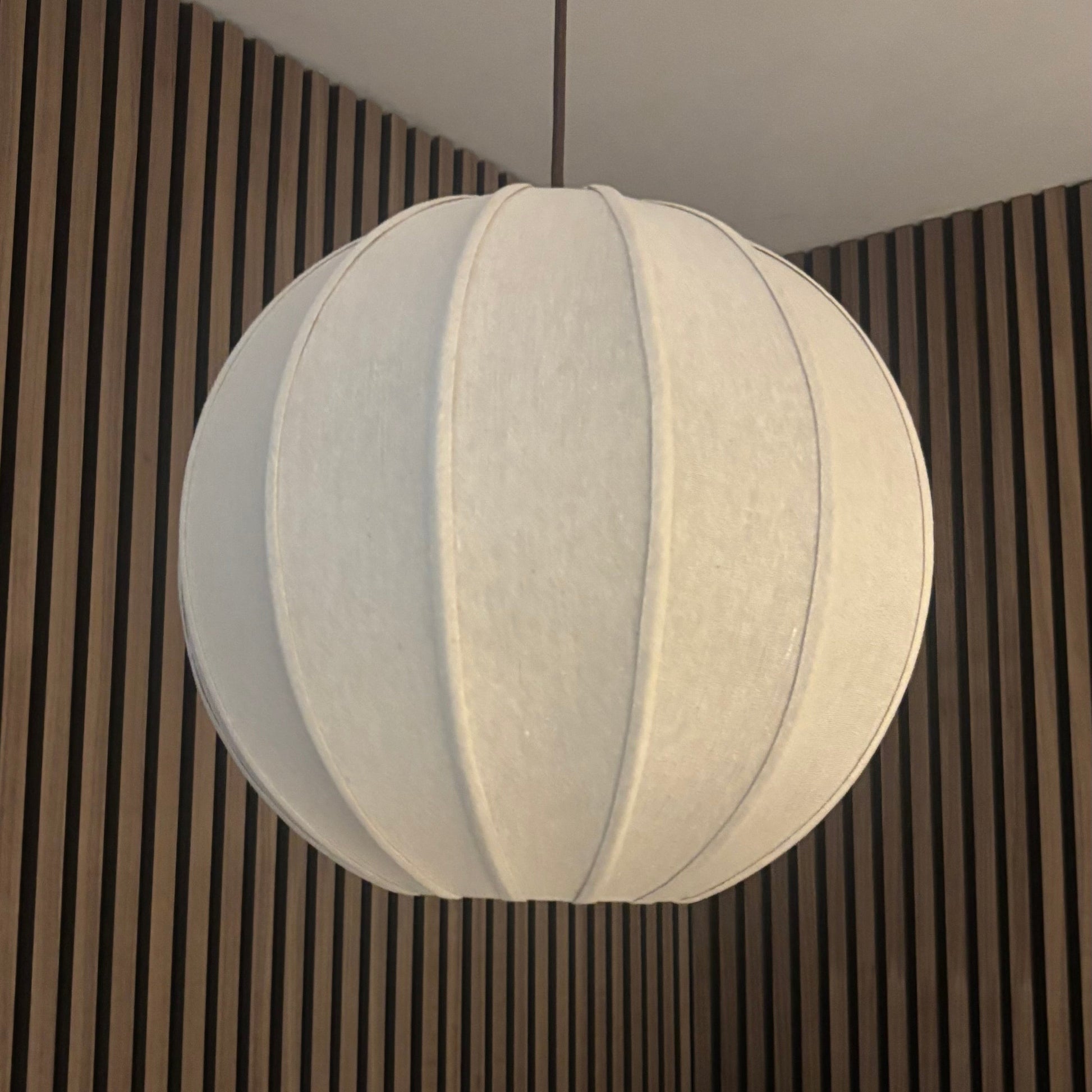 A white fabric paper lantern pendant light hanging from a black cord in a room with brown vertical wood slats and a green plant in the background.