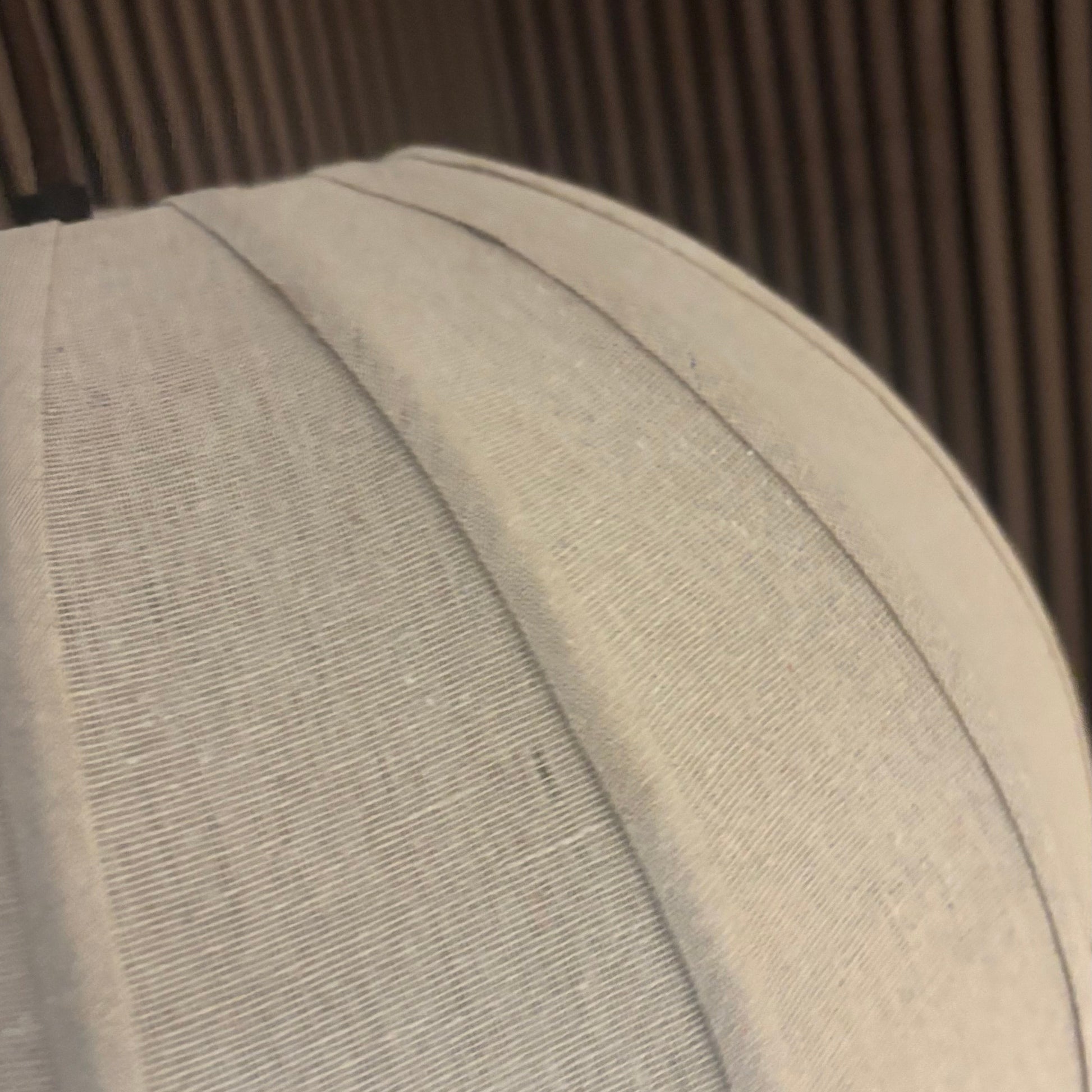 A close-up of a light beige or off-white textured fabric dome, likely a display stand or dome cap, set against a dark brown slatted background.