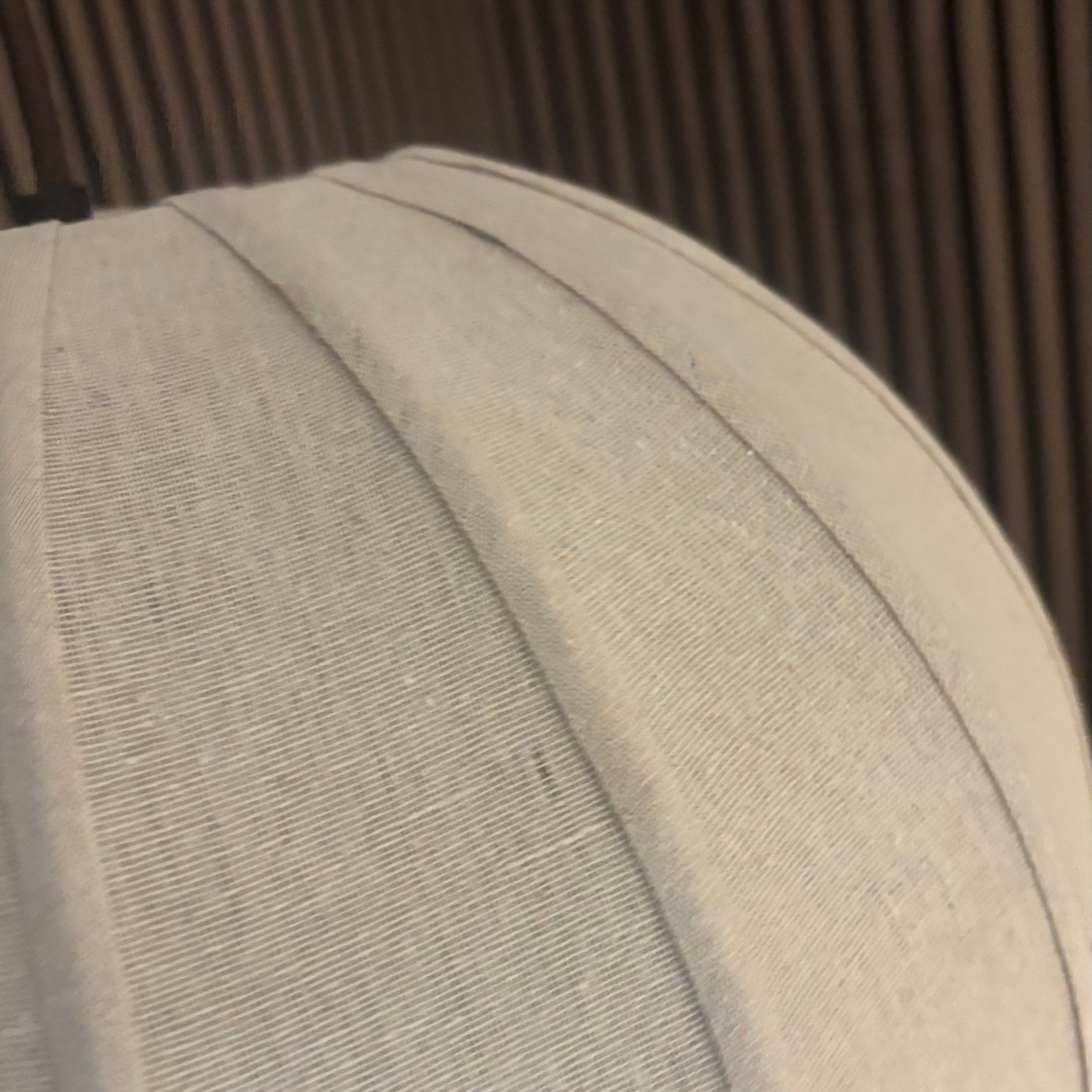 A close-up of a light beige or off-white textured fabric dome, likely a display stand or dome cap, set against a dark brown slatted background.
