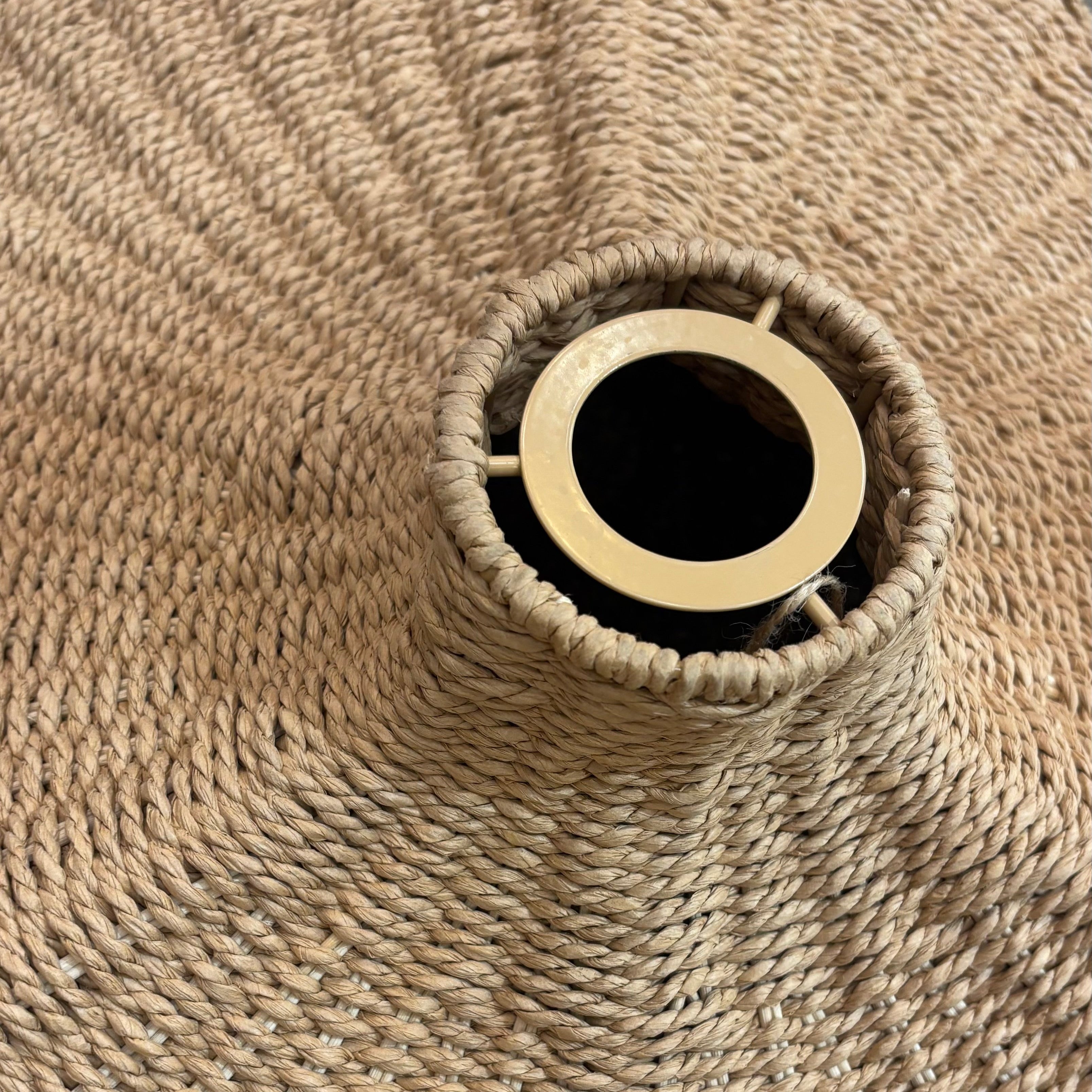A close-up of a woven natural fiber hat, likely a straw sun hat or wide-brimmed hat, featuring a circular opening at the top with a light wood or brass rim.