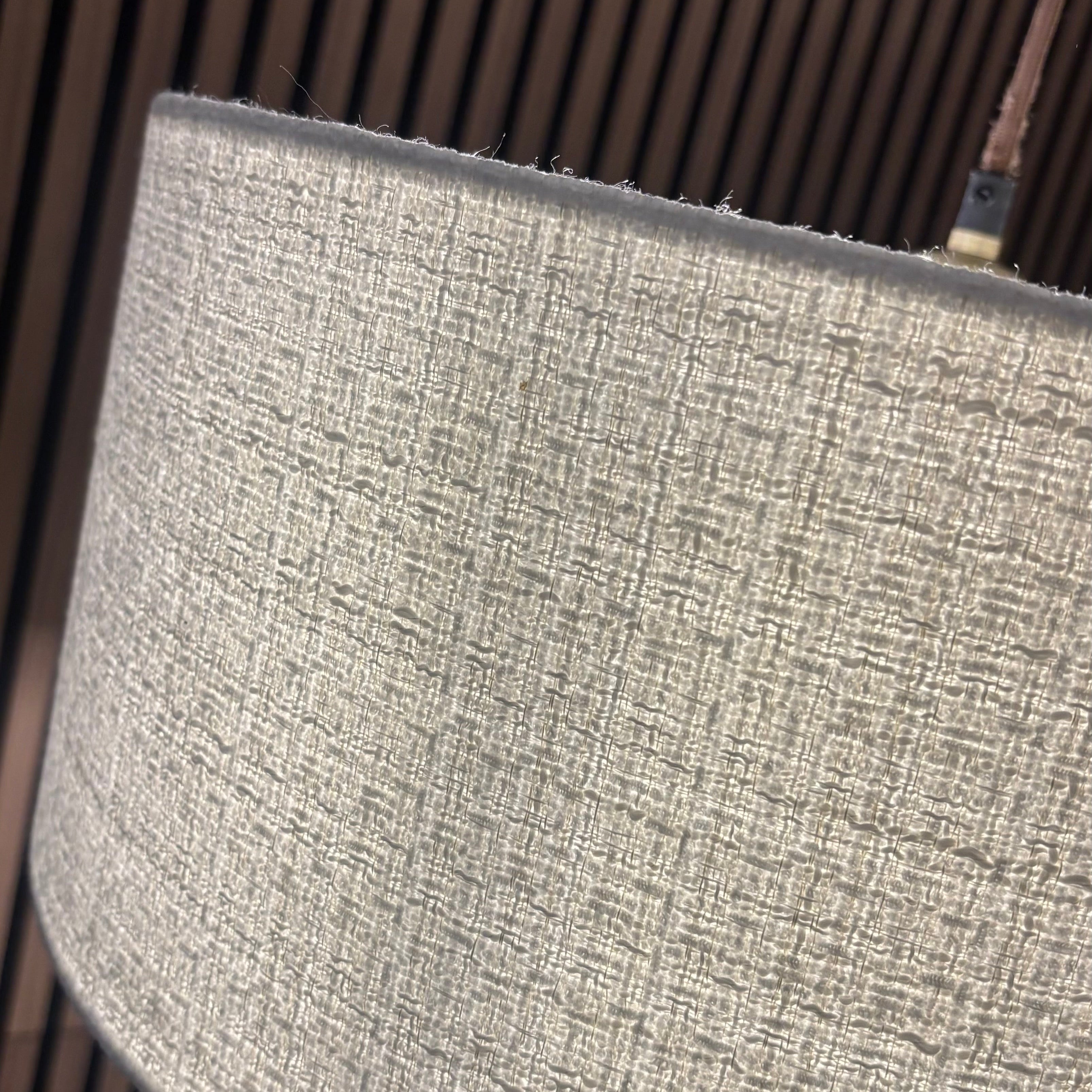 Close-up of a cylindrical lampshade made of textured woven fabric in a light gray color, set against a dark brown slatted background.