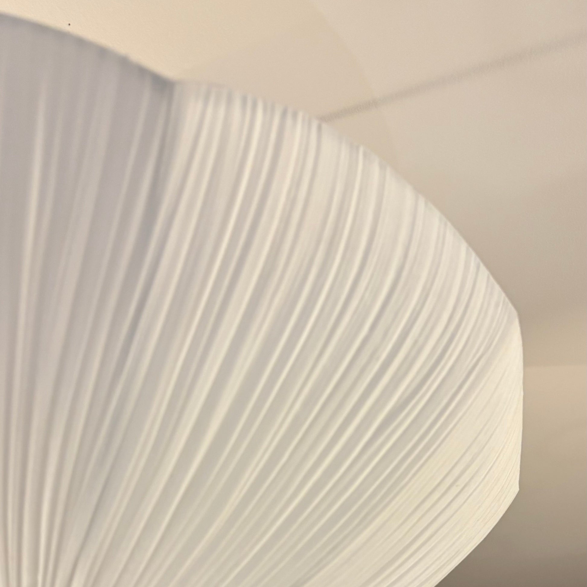 A close-up of a modern pleated white ceiling light fixture, showing its textured fabric shade against a dark wood slatted wall and white ceiling.