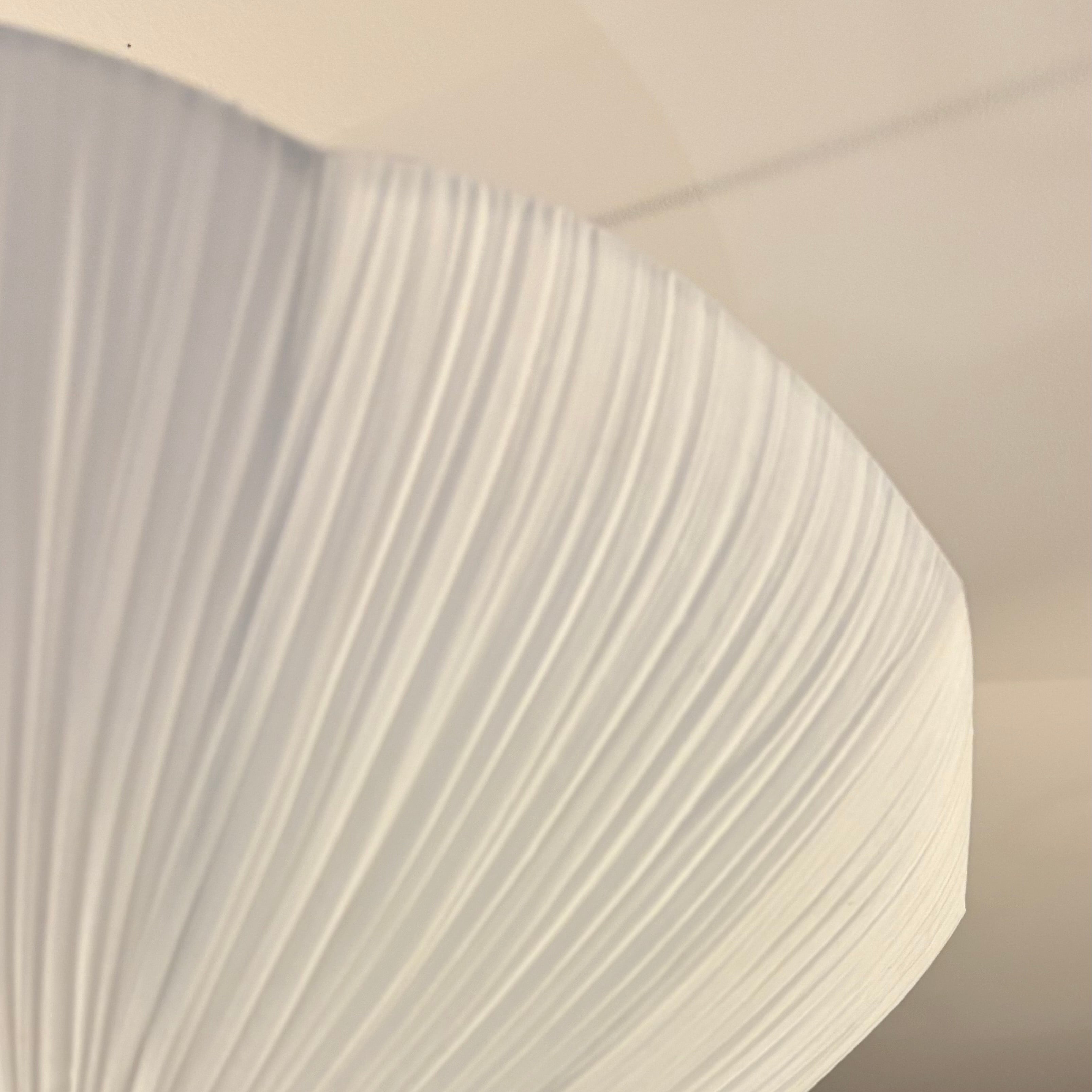 A close-up of a modern pleated white ceiling light fixture, showing its textured fabric shade against a dark wood slatted wall and white ceiling.