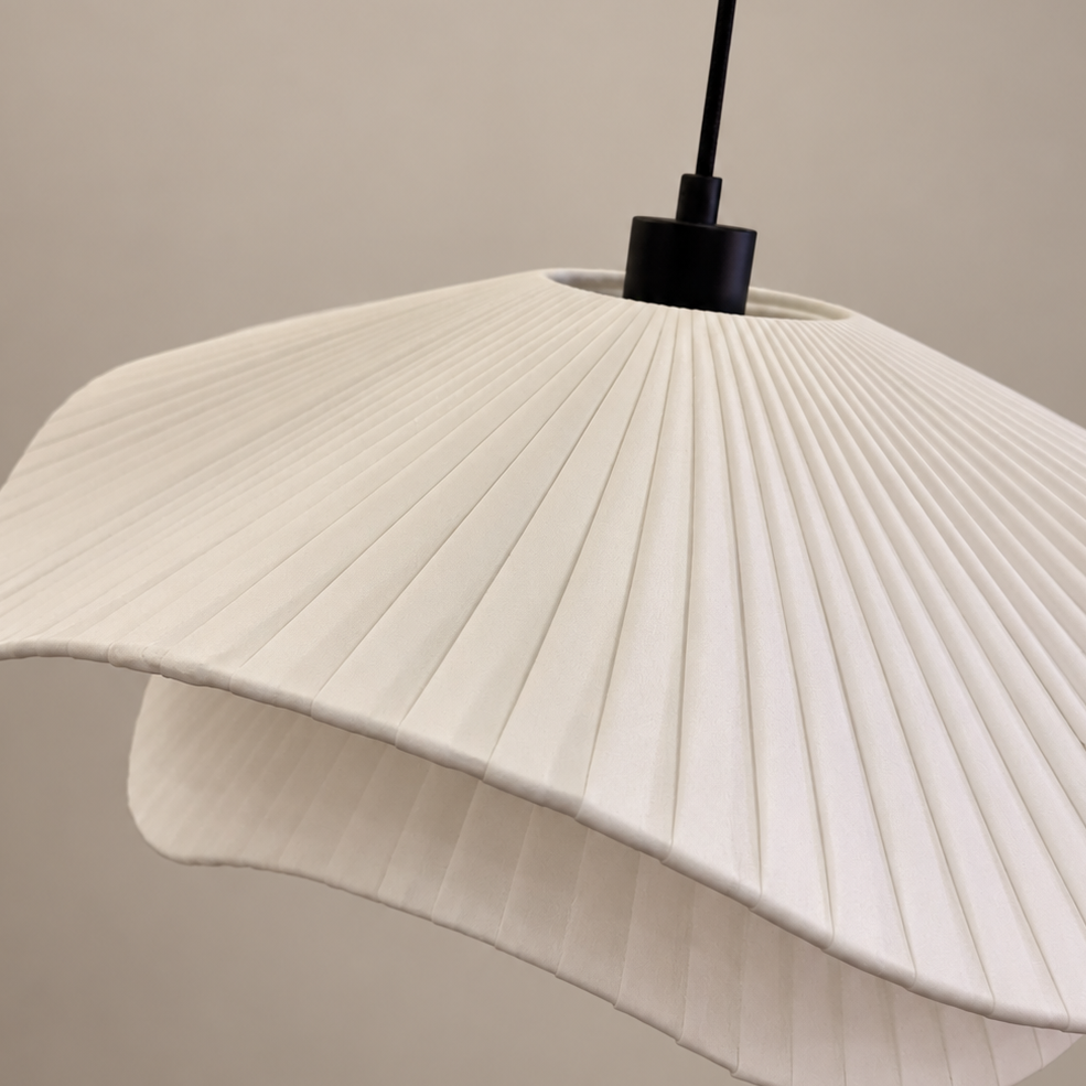 A close-up of a modern pleated pendant light shade in off-white, suspended from a black ceiling canopy by a black cord against a warm beige background.