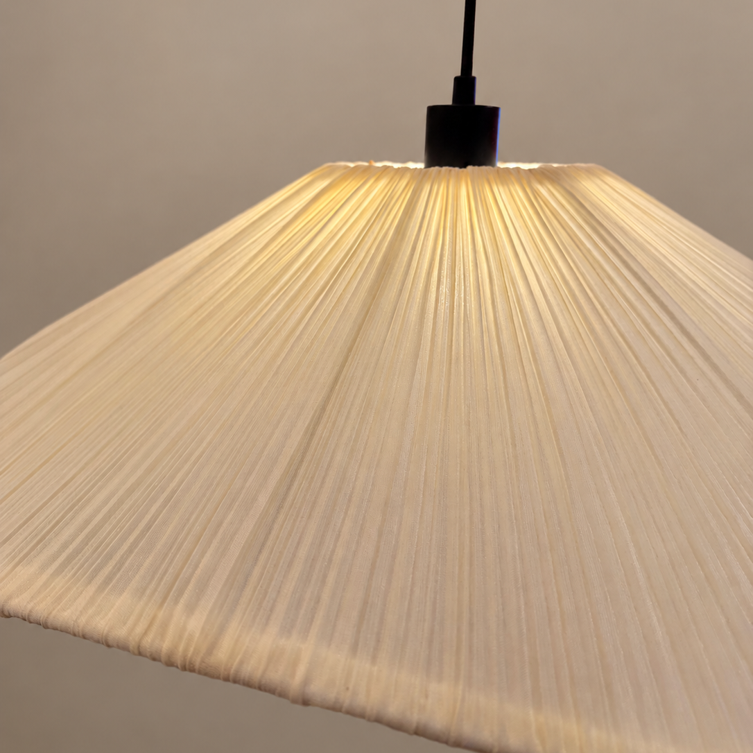 A close-up of a pleated fabric pendant lamp shade glowing warm white, suspended from a black cord against a plain beige wall.