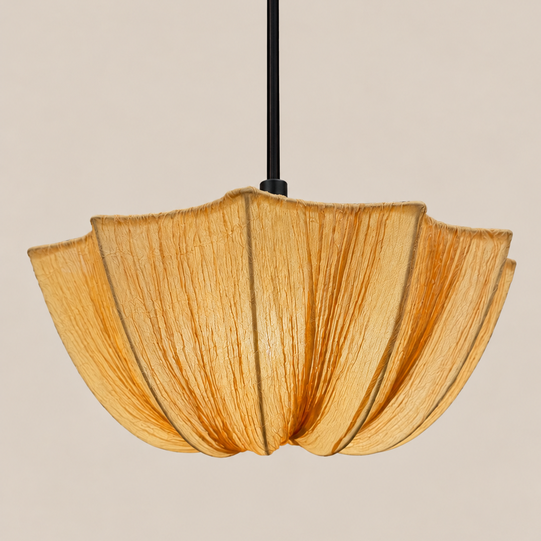 A modern pendant light with a textured, pleated shade made of natural woven fibers in a warm golden-brown hue, suspended by a slim black cord against a solid light beige background.