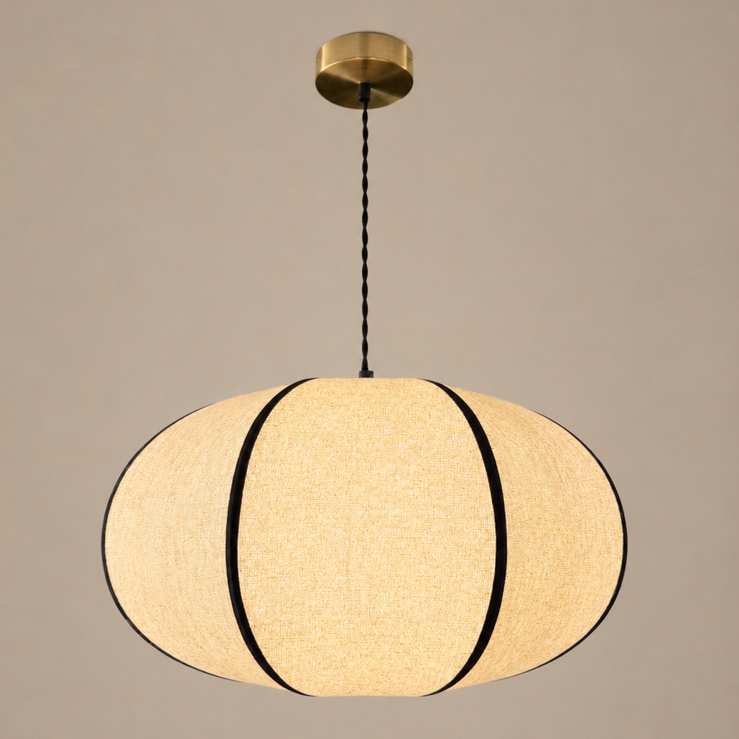 A modern pendant light with a textured beige fabric shade, accented by black vertical bands and a gold brass ceiling canopy, shown against a solid warm gray background.