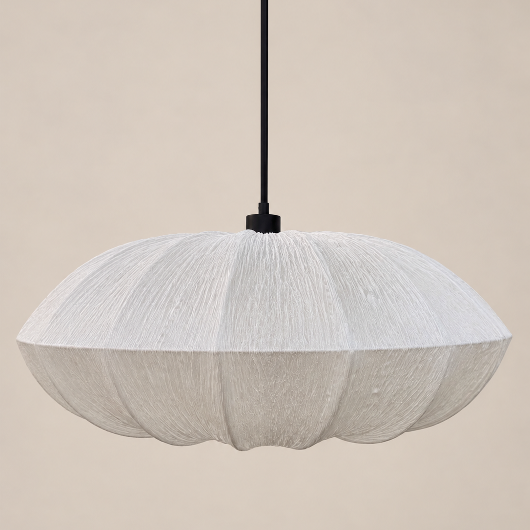 A modern pendant light with a textured, pleated white shade in a scalloped, flower-like shape, suspended by a thin black cord against a solid beige background.